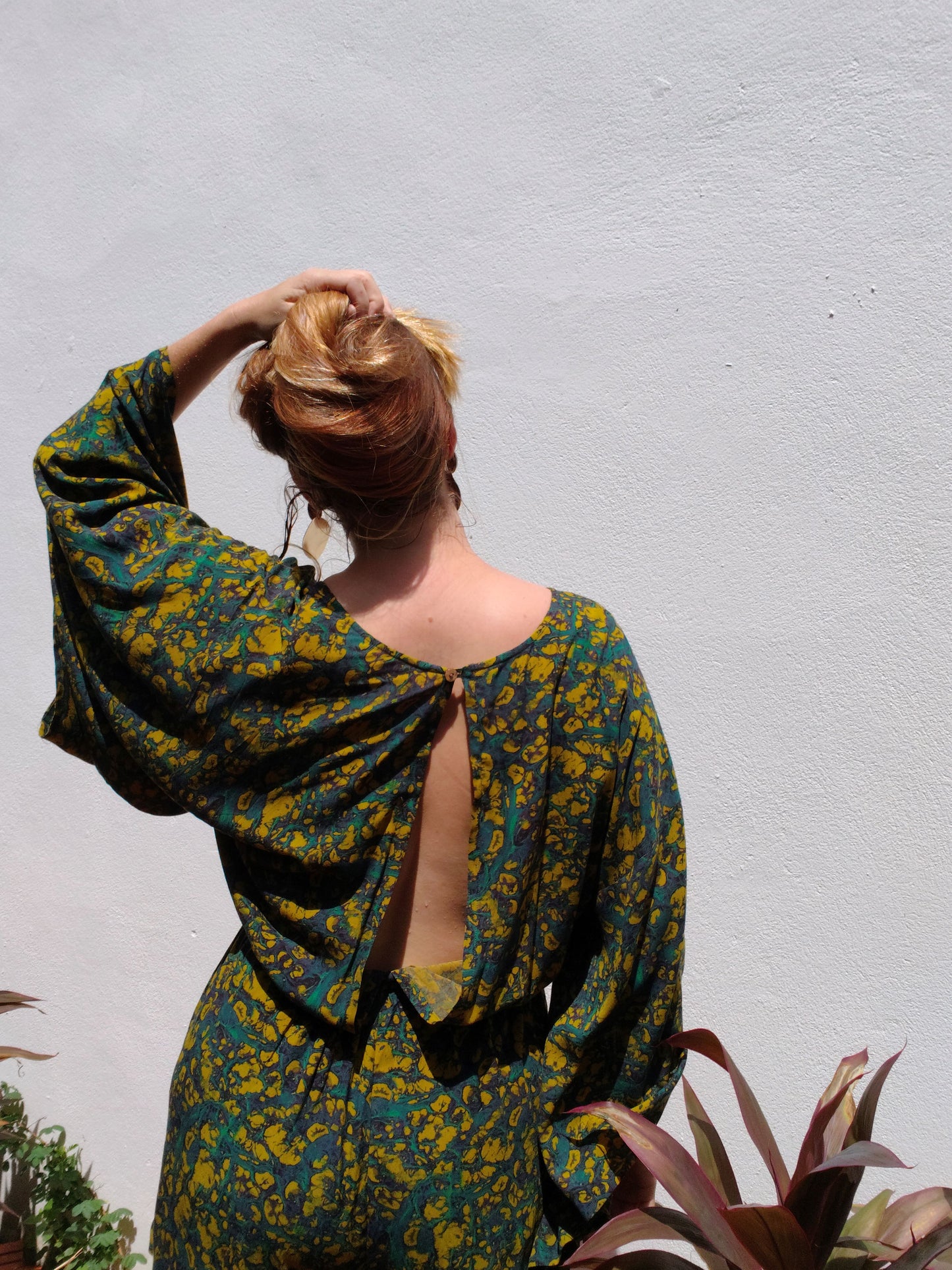 Woman wearing a green and yellow patterned romper with a backless design, standing against a light gray wall.