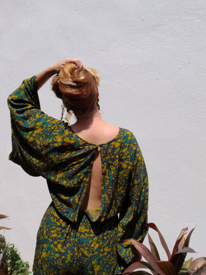Woman wearing a green and yellow patterned romper with a backless design, standing against a light gray wall.