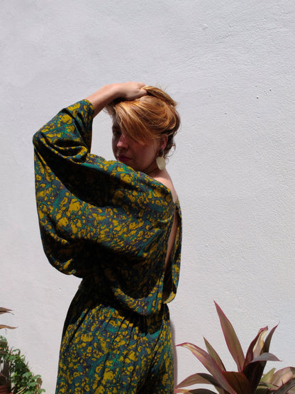 Person wearing a green and yellow patterned outfit against a white wall with plants.